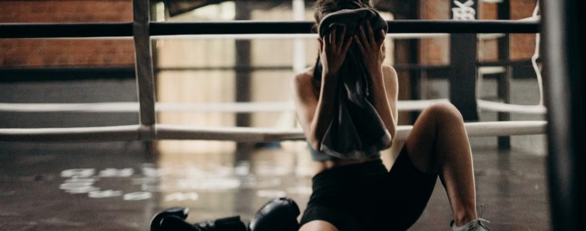 A female boxer sitting in a gym ring, resting and wiping sweat with a towel after a workout.